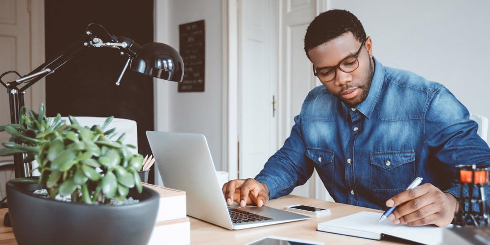 A man works at a laptop while writing in a notebook, with a bicycle in the background.