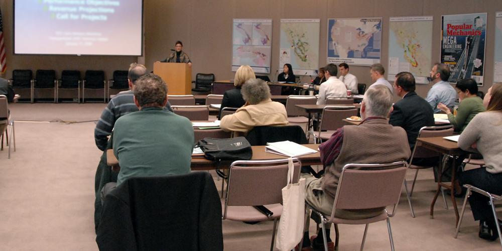 Adults in a classroom setting, all looking forward at a woman presenting a slideshow at the front of the room.