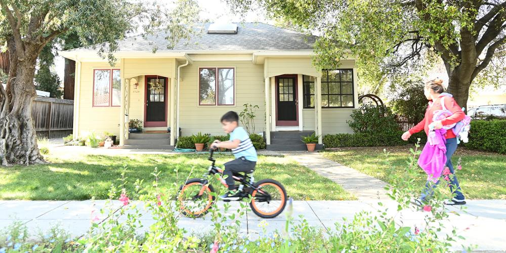 A child on a bicycle and their parent walking on a sidewalk in front of a single-family home.