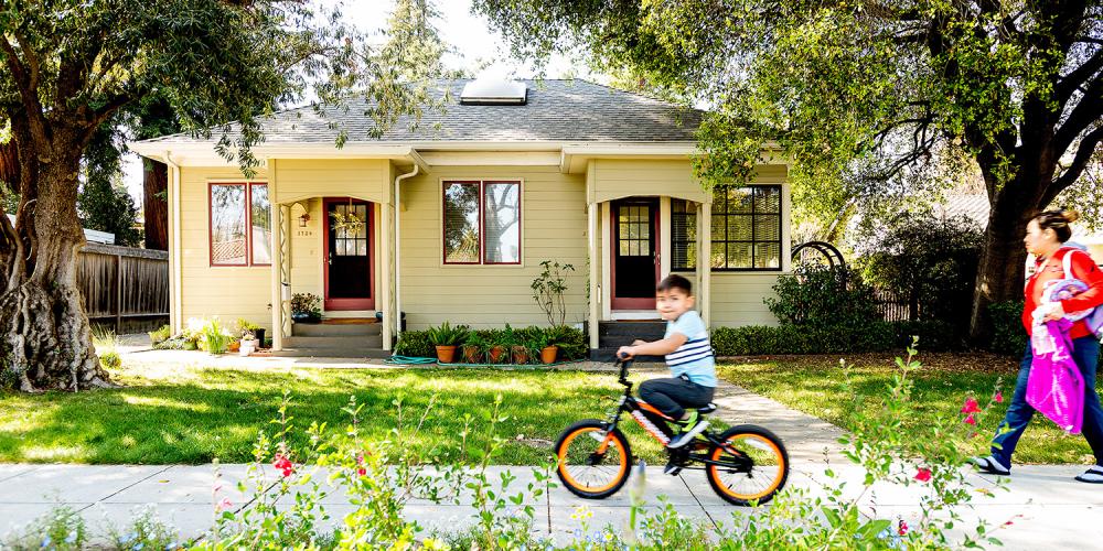 A boy on a bicycle and his parent on the sidewalk passing a home in the South Bay.