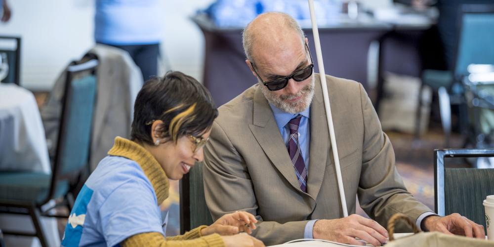 Latino woman in blue shirt sits with visually impared man wearing sunglasses, woman is examining a document  