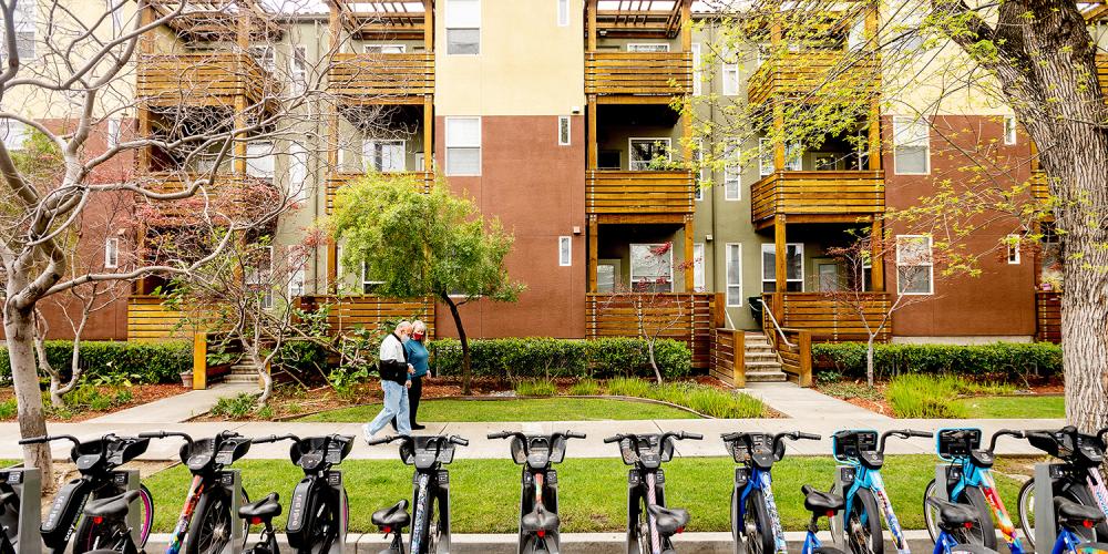An apartment complex with a bike share rack in the foreground and adults walking on the sidewalk.