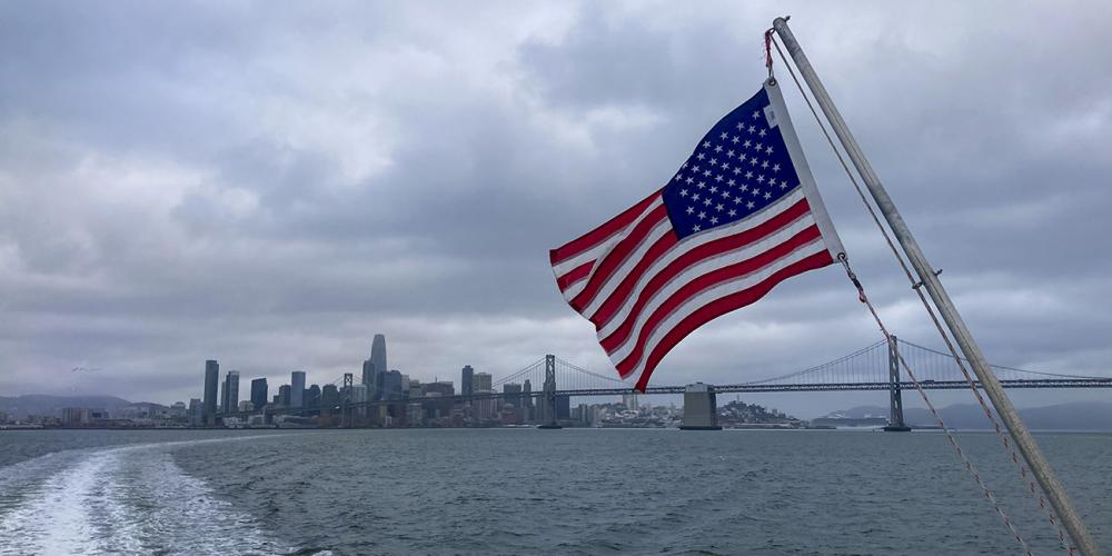 An American flag with the San Francisco-Oakland Bay Bridge and San Francisco skyline in the background.