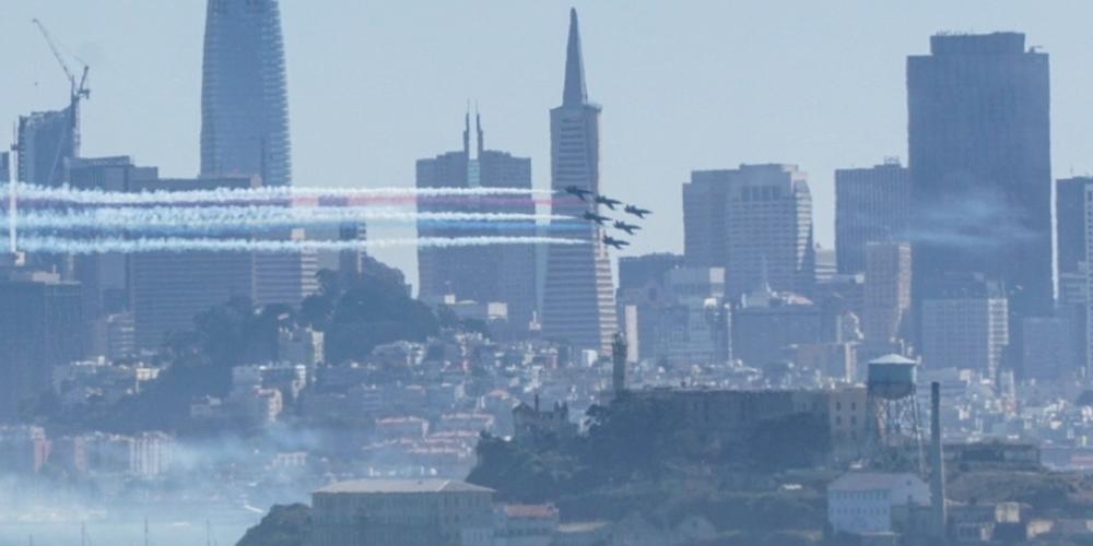 SF Skyline with blue angels flying by with red white and blue smoke. 