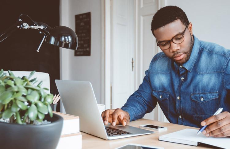 A man works at a laptop while writing in a notebook, with a bicycle in the background.