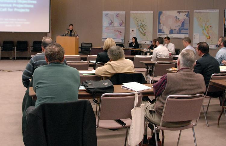 Adults in a classroom setting, all looking forward at a woman presenting a slideshow at the front of the room.