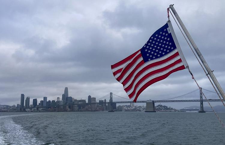 An American flag with the San Francisco-Oakland Bay Bridge and San Francisco skyline in the background.